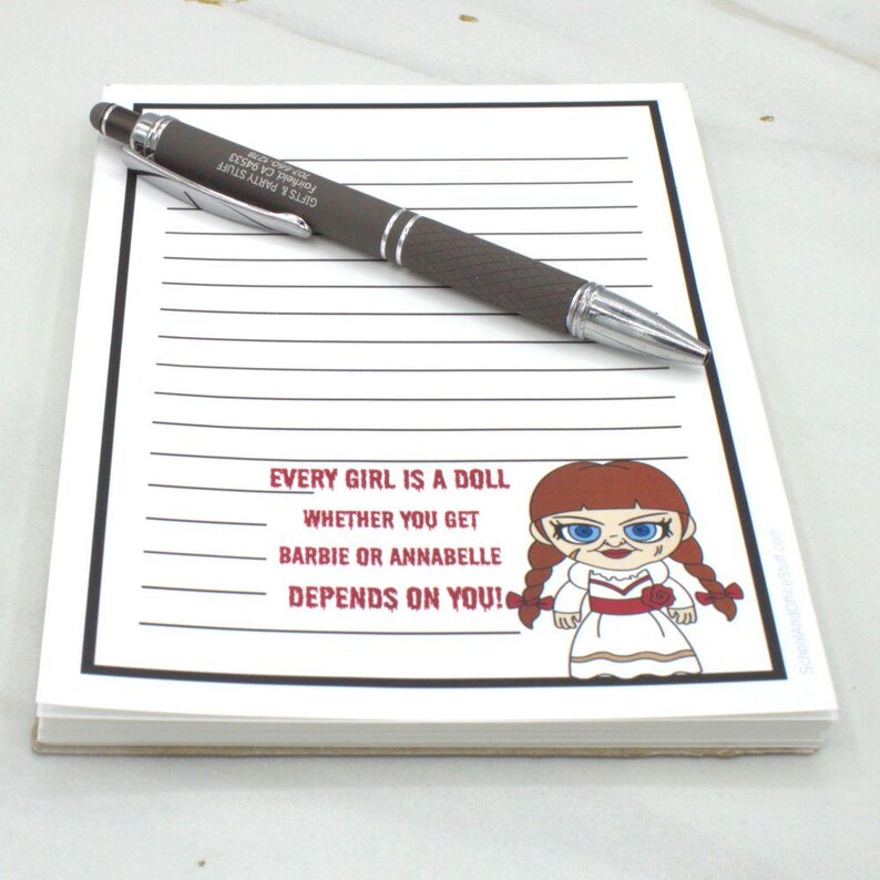 May include: A notepad with lined paper and a dark brown pen. The notepad has the text "EVERY GIRL IS A DOLL WHETHER YOU GET BARBIE OR ANNABELLE DEPENDS ON YOU!" and a cartoon doll illustration.