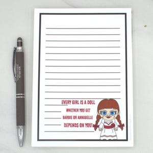 May include: A lined notepad with a black border and the text "EVERY GIRL IS A DOLL, WHETHER YOU GET BARBIE OR ANNABELLE DEPENDS ON YOU!" next to a cartoon doll. A brown and silver pen is to the left.