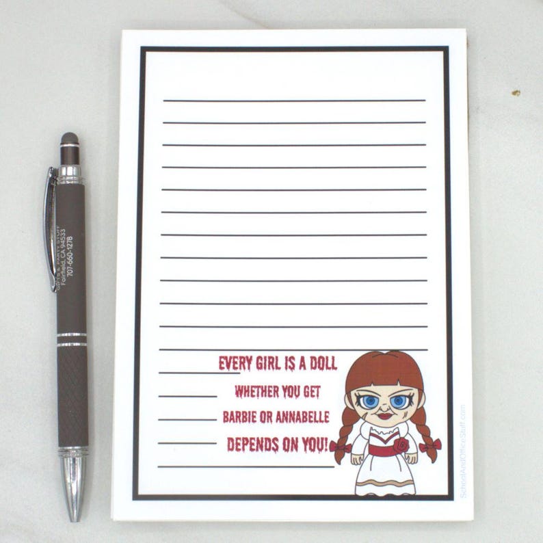 May include: A lined notepad with a black border and the text "EVERY GIRL IS A DOLL, WHETHER YOU GET BARBIE OR ANNABELLE DEPENDS ON YOU!" next to a cartoon doll. A brown and silver pen is to the left.