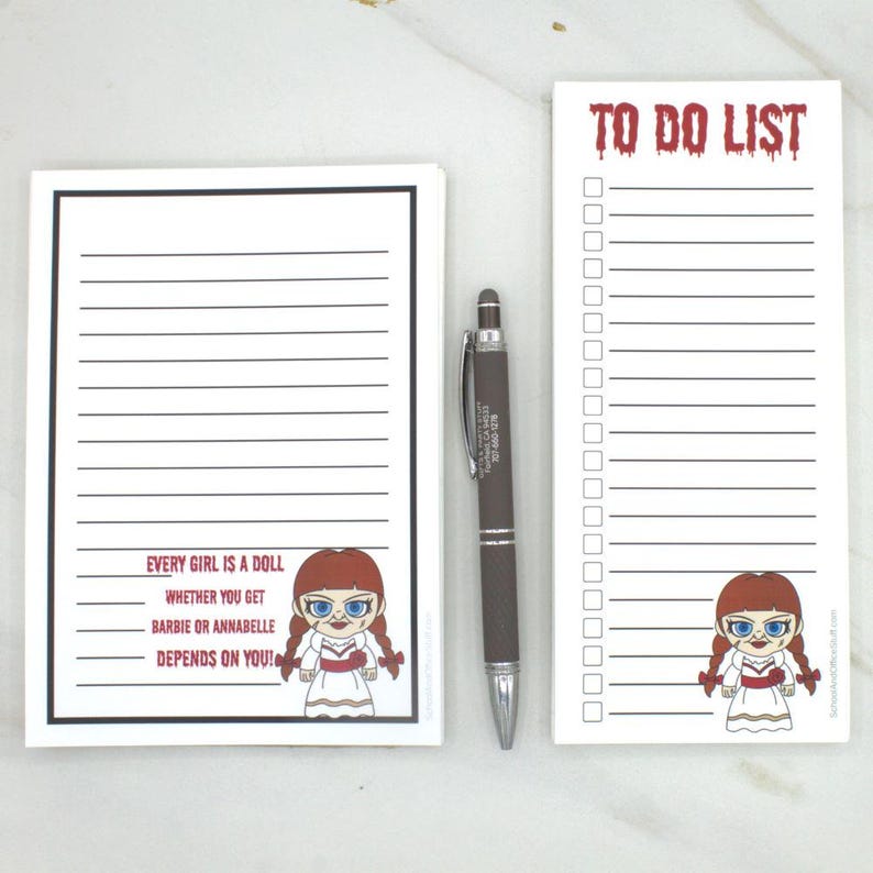 May include: Two white notepads with black lines and text. One notepad says "EVERY GIRL IS A DOLL WHETHER YOU GET BARBIE OR ANNABELLE DEPENDS ON YOU!" with a doll graphic. The other says "TO DO LIST" with a doll graphic. A pen is between the notepads.