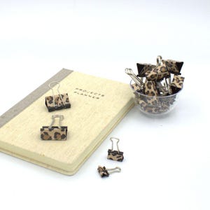May include: A beige Projects Planner notebook with a gray spine, accompanied by several metal binder clips featuring a brown and black leopard print pattern. Some clips are placed on the notebook cover, while others are in a clear bowl.