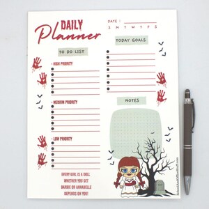 May include: A white daily planner with a spooky theme. The planner features sections for to-do lists, notes, and today's goals. Red handprints and bats decorate the page. A brown pen rests beside the planner. The text reads "Daily Planner".