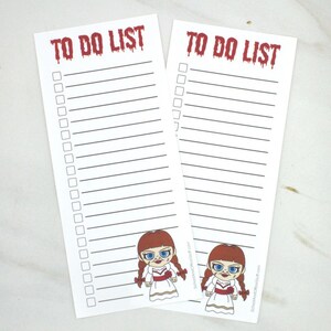 May include: Two white "To Do List" notepads with a red, dripping font. Each notepad has a lined section for writing and a cartoon image of a doll at the bottom. The doll has red hair, blue eyes, and a white dress.