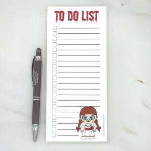 May include: A white To Do List notepad with a cartoon Annabelle doll illustration and a dark gray pen. The notepad has a red, dripping font that reads "TO DO LIST". The pen is next to the notepad.