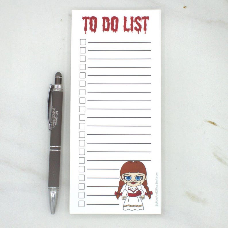 May include: A white To Do List notepad with a cartoon Annabelle doll illustration and a dark gray pen. The notepad has a red, dripping font that reads "TO DO LIST". The pen is next to the notepad.