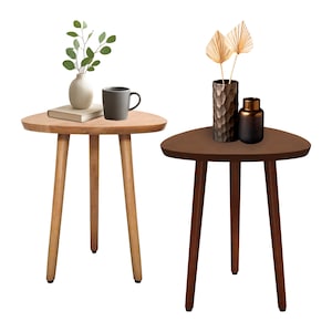 May include: Two triangular side tables with three legs each. One table is light wood with a vase, book, and mug. The other is dark wood with a textured vase and a small, dark bottle. The tables are on a white background.