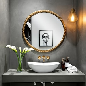 May include: A modern bathroom vanity with a large, round gold-framed mirror. A white oval sink sits on a gray countertop, with gold fixtures and a vase of white calla lilies. A framed portrait hangs on the wall.