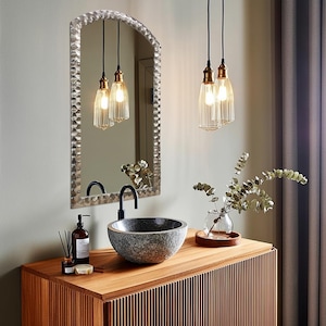 May include: A bathroom vanity with a wooden cabinet, a stone sink, and a black faucet. Above the sink is a textured, arched mirror. Two pendant lights hang above the sink, and a vase of greenery sits on the counter. The overall color scheme is neutral with warm wood tones.