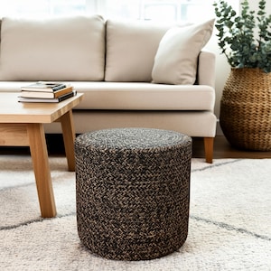 May include: A cylindrical ottoman with a woven pattern in shades of brown and black. The ottoman is placed on a white rug with a geometric design, near a light-colored sofa and wooden coffee table. A wicker basket with greenery is in the background.