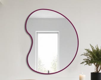 May include: A modern, irregularly shaped mirror with a deep burgundy frame hangs on a white wall. Below, a black table holds a brown bowl, a white candle, and a potted plant. The mirror reflects a window.