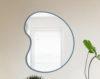 May include: An irregularly shaped mirror with a blue frame hangs on a cream-colored wall. Below, a black table displays a green potted succulent, a small gray vase, and a plant in a beige pot. The mirror reflects a window with white blinds.