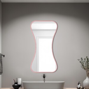 May include: A pink-framed, hourglass-shaped mirror hangs above a gray oval sink in a modern bathroom. The sink sits on a light wood shelf with towels and decorative items. A black faucet and towel rack are also visible.