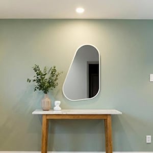 Op de afbeelding: Een moderne entree met een lichtgroene muur, een houten console met een wit blad en een unieke, asymmetrische spiegel. Een vaas met groen en een decoratief object staan op de tafel. De kamer heeft hardhouten vloeren en een neutraal tapijt.