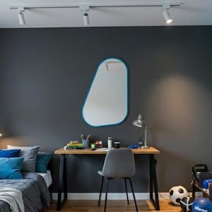 Op de afbeelding: Een moderne slaapkamer met een bed, bureau en spiegel. Het bed heeft blauwe en grijze beddengoed met decoratieve kussens. Een houten bureau met een grijze stoel staat voor een donkergrijze muur. Een blauw omrande spiegel hangt aan de muur. De kamer heeft houten vloeren.