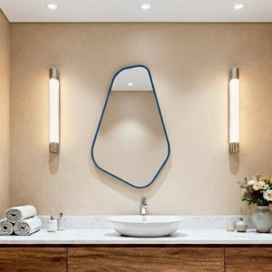 May include: A modern bathroom vanity with a wooden cabinet and a white countertop. A unique, blue-framed mirror hangs above a white, oval sink. Two vertical wall sconces flank the mirror. Rolled white towels and a vase of flowers add decorative touches.