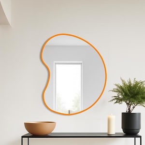 May include: An orange-framed, organically shaped mirror hangs above a black console table. A small potted plant, a candle, and a wooden bowl are on the table. A tall, slender lamp stands to the left.