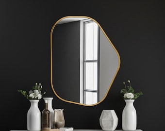May include: A gold-framed, irregularly shaped mirror hangs on a black wall. Below, a white shelf displays white vases with flowers, a brown bottle, and a geometric vase. The scene is minimalist and modern.