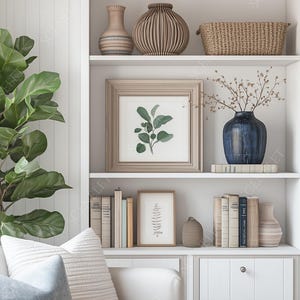 May include: A white built-in bookshelf with decorative items. It features framed botanical prints, a blue vase with branches, books, and various ceramic vases. A large green plant is to the left of the bookshelf.