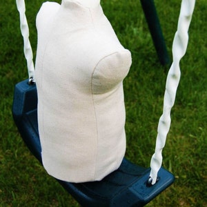 May include: A white fabric torso form is hanging from a swing set. The swing set is made of metal and has a blue plastic seat. The background is a green grassy lawn.