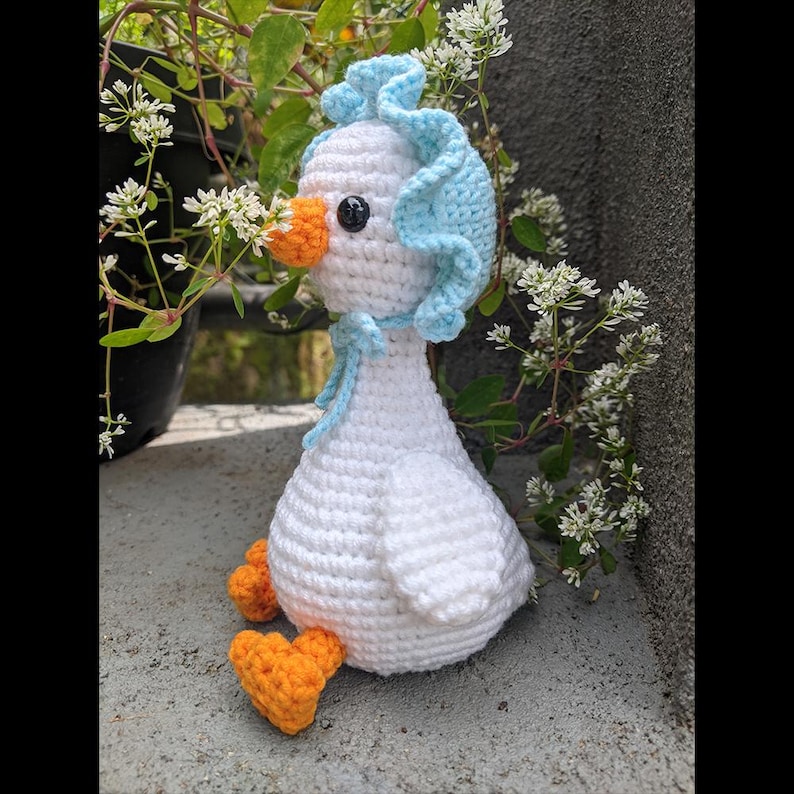 May include: A handmade crochet goose with a white body, orange beak and feet, and a light blue bonnet. The goose has a black eye and a blue ribbon tied around its neck. The crochet goose is sitting on a concrete surface.
