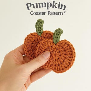 May include: Two orange crochet pumpkin coasters with green stems are held in a hand. The coasters are round and have a textured appearance. The text "Pumpkin Coaster Pattern" is at the top of the image.