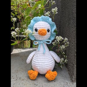 May include: A handmade crochet duckling doll. The duckling is white with an orange beak and feet. It wears a light blue bonnet tied with a matching bow. The doll is sitting on a concrete surface with greenery in the background.