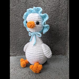May include: A handmade crochet duckling doll. The duckling is primarily white with a light blue bonnet and bow. It has an orange beak and feet, and black eyes. The doll is sitting on a gray surface.