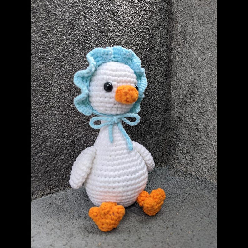 May include: A handmade crochet goose doll with a white body, orange feet and beak, and a light blue bonnet tied with a bow. The doll is sitting on a gray surface.