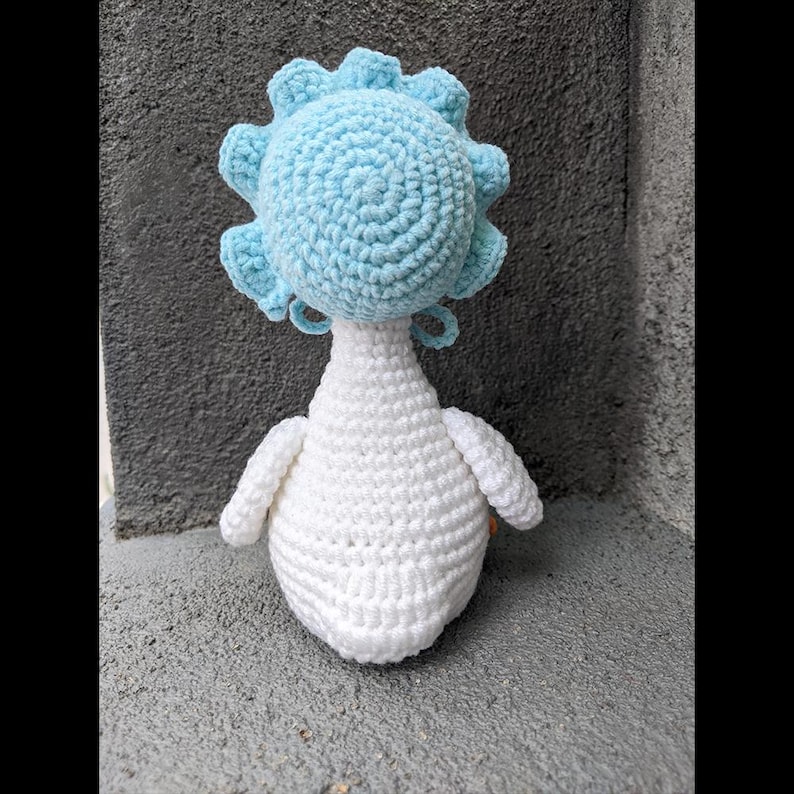May include: A handmade crochet doll with a light blue head and white body. The head has a circular shape with a ruffled edge. The body is pear-shaped with small, rounded wings. The doll is made of soft yarn.