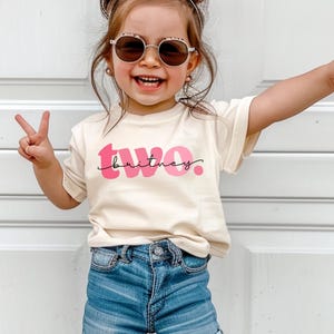 May include: A cream-colored short-sleeve t-shirt featuring the word two in large pink letters with the name Britney written in black cursive script across the center. The shirt is paired with blue denim shorts, round sunglasses, and a pink hair bow.