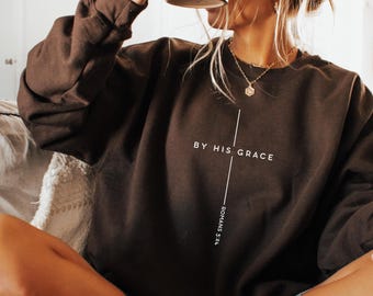 May include: Dark chocolate-colored sweatshirt with the text "BY HIS GRACE" and "ROMANS 5:24" printed in white. The sweatshirt has a crew neck and long sleeves. The image also includes a black mug.