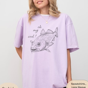 May include: A lilac t-shirt featuring a detailed illustration of a fish and the pun "oh my cod!" in black script. The shirt has a loose fit and short sleeves. The image also includes a speech bubble that says "Sweatshirts, Long Sleeve Available".