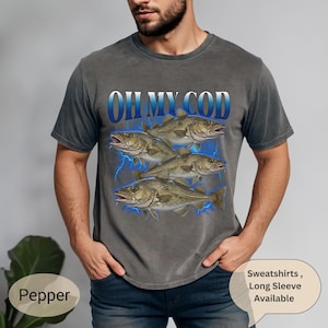 May include: Dark grey t-shirt with the text "OH MY COD" in white and blue, and a graphic of four fish with blue lightning. A casual, comfortable style of shirt.