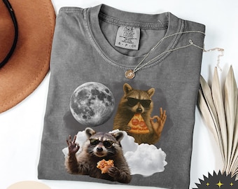 May include: A gray Comfort Colors t-shirt featuring a graphic of two raccoons wearing sunglasses, with a pizza slice and a donut. A full moon and clouds are also in the design. A silver necklace is draped across the shirt.