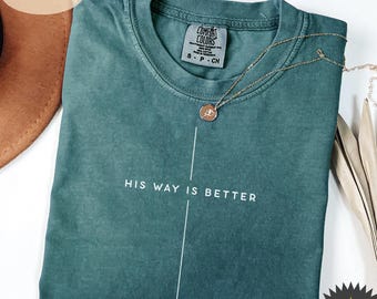 May include: Blue spruce t-shirt with the text "HIS WAY IS BETTER" in white. A gold necklace with a pendant is draped over the shirt. The shirt is folded and has a tag that says "Comfort Colors".