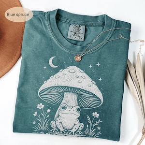 May include: A blue spruce colored t-shirt featuring a white graphic print of a frog sitting under a large mushroom with a crescent moon and stars in the background. The shirt has a Comfort Colors label at the neck and a thin gold chain necklace.