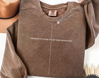 May include: Espresso-colored Comfort Colors sweatshirt with the text "HEAVEN IS MY HOME I'M JUST HERE RECRUITING" in white. A silver necklace with a small pendant is draped across the front. A brown hat is visible in the background.