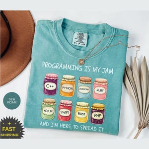 May include: Seafoam green t-shirt featuring the phrase "PROGRAMMING IS MY JAM" and jam jar illustrations representing programming languages such as C++, Python, and Ruby. The shirt also has the text "AND I'M HERE TO SPREAD IT".