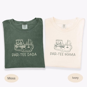 May include: Two t-shirts, one moss green and one ivory, are displayed. Each shirt features a golf-themed graphic with a golf cart, flag, and the words "PAR-TEE DADA" and "PAR-TEE MAMA" in white. The shirts are folded.