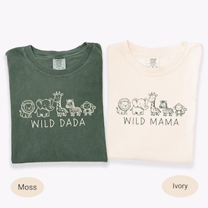 Op de afbeelding: Twee T-shirts, één mosgroen en één ivoor, elk met een ronde hals. Elk heeft de woorden "WILD DADA" en "WILD MAMA" gedrukt onder een rij cartoon dieren. De shirts zijn gemaakt van een zacht materiaal.