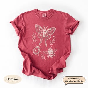 Op de afbeelding: Een karmozijnrood t-shirt met korte mouwen en een witte lijntekening van een grote nachtvlinder, een lieveheersbeestje en een bij, omringd door kleine takjes met blaadjes. Het t-shirt heeft een ronde halslijn.