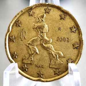 Może przedstawiać: Złota moneta z wystylizowanym wizerunkiem postaci i rokiem 2002. Moneta ma szereg gwiazd na obrzeżu, widoczne są litery "R" i "MAC". Moneta jest trzymana w przezroczystym plastikowym uchwycie.