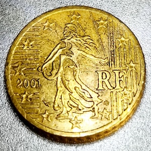 May include: A gold-coloured coin with intricate detailing. The coin depicts a woman in robes, stars, and the year 2001. The letters "RF" are also present. This is likely a Euro coin.