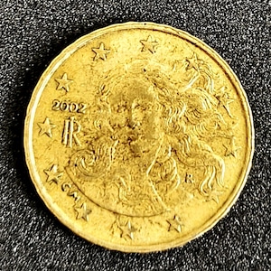 May include: A gold-coloured coin with a woman's profile, stars, and the year 2002. The coin has the letters "IR" and other inscriptions. The coin is circular and has a slightly worn appearance.