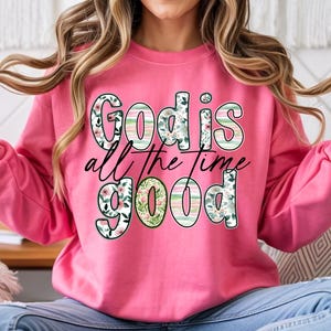 Puede incluir: Una sudadera rosa con el texto "God is all the time good" en una mezcla de patrones florales y a rayas. La sudadera es de color rosa liso.