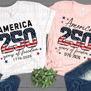 May include: A white t-shirt and a light pink t-shirt, each featuring the text AMERICA 250 years of freedom 1776-2026. The number 250 is printed with a red, white, and blue American flag pattern, accompanied by small red and blue star graphics.