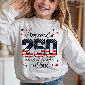 May include: A white long-sleeve sweatshirt featuring a graphic with the text America 250 years of freedom 1776-2026. The number 250 is filled with a red, white, and blue American flag pattern, surrounded by small red and blue stars.