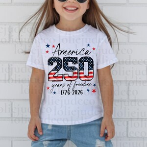 May include: A white short-sleeve t-shirt featuring a graphic with the text America 250 years of freedom 1776-2026. The numbers 250 are filled with a red, white, and blue American flag pattern, surrounded by small red and blue stars.