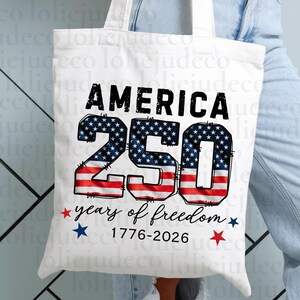 May include: A white canvas tote bag featuring the black text AMERICA above the large number 250, which is filled with a red, white, and blue American flag pattern. Below, the text reads years of freedom 1776-2026 with small red and blue stars.