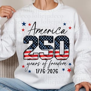 May include: A white long-sleeve sweatshirt featuring a graphic with the text America 250 years of freedom 1776-2026. The number 250 is filled with a red, white, and blue American flag pattern, surrounded by small red and blue stars.